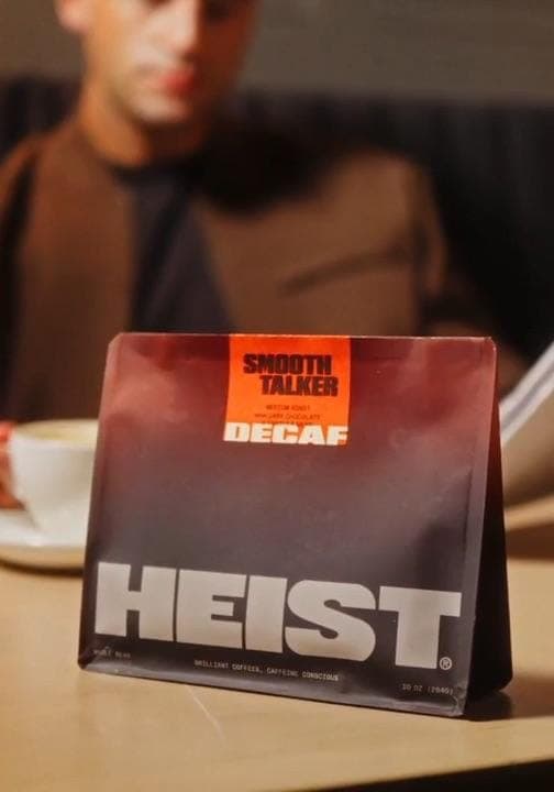 The image shows a bag of "Smooth Talker Decaf" coffee beans resting on a wooden table. The bag is primarily dark red/brown with a gradient effect, and features the brand name "Heist" in white letters. The bag also includes the text "Smooth Talker Decaf" and "Brilliant Coffee, Caffeine Connoisseurs" in smaller white let