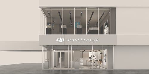 A rendered exterior view of a two-story storefront with a large glass facade and a light grey sign band reading "DJI | HASSELBLAD". Through the glass, the interior shows display counters, product shelves, and a tall digital screen on the right.