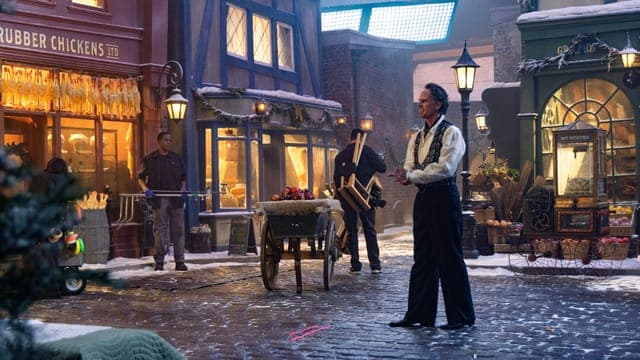 The image depicts a man in a black suit and white shirt standing on a cobblestone street, holding a wooden stick or rod. He is positioned in front of a store with a sign that reads "Rubber Chickens". The store is decorated with Christmas lights and garland, and a few people are visible in the background. The street is