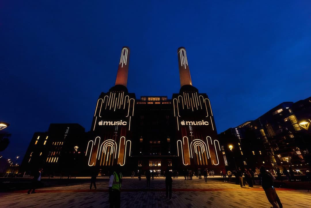 Battersea Power Station at night, with its chimneys and facade outlined in white neon-style artwork and "music" Apple Music logos on the walls. Pedestrians stand in the plaza in front of the building under a deep blue sky.