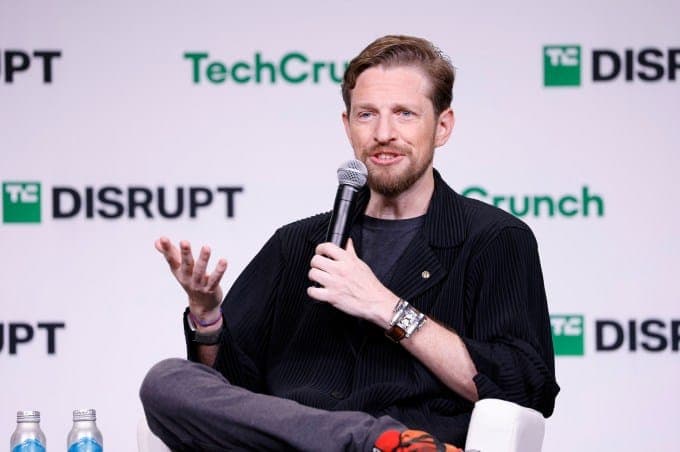 The image shows a man seated on a white chair, holding a microphone and gesturing with his right hand. He is wearing a black jacket over a dark-colored t-shirt and has a beard. He is also wearing a watch on his left wrist. The backdrop is a white wall with the logos of TechCrunch and Disrupt, suggesting a tech-related