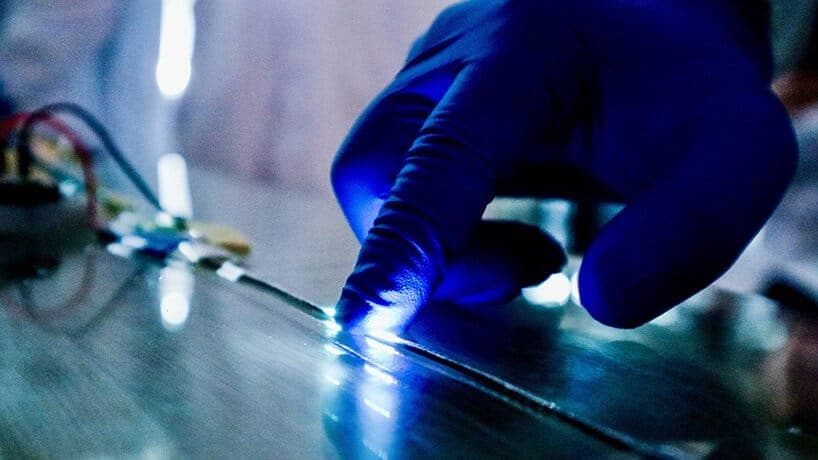 A gloved hand, wearing a vibrant blue glove, is delicately pressing a small, bright blue light onto a metallic surface. The light is reflected on the surface, creating a mesmerizing pattern of blue hues. The background is blurred, drawing focus to the hand and the light source. The metallic surface appears to be a flat