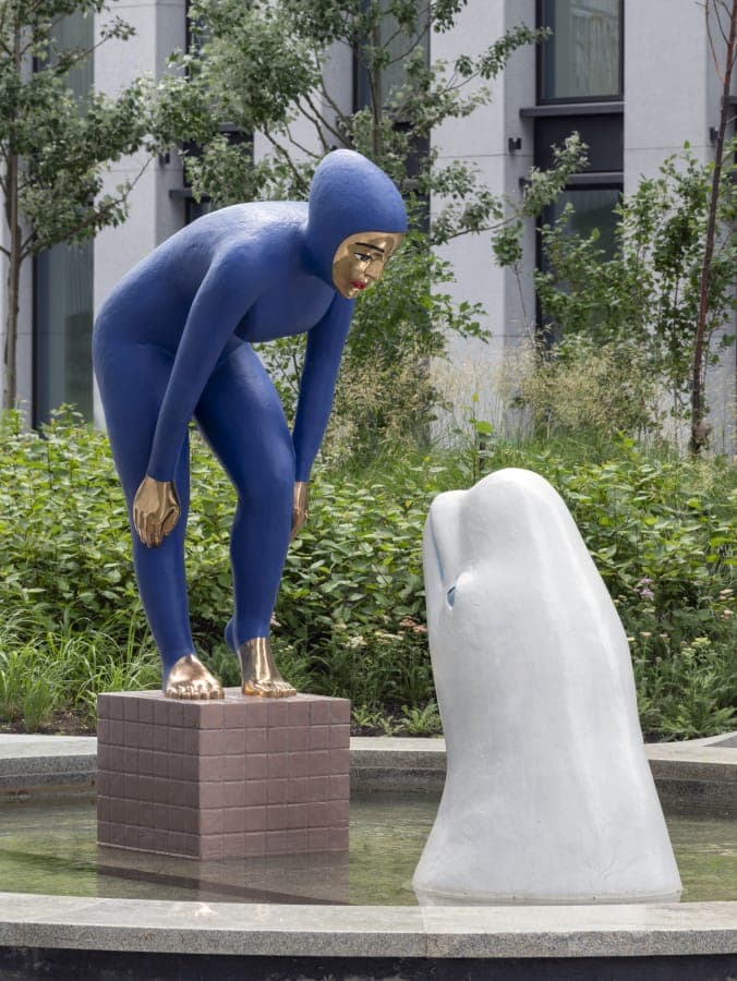 The image depicts a striking sculpture of a person in a blue bodysuit kneeling on a brick pedestal, facing a white dolphin. The dolphin is positioned on the right side of the pedestal, with its head turned towards the person. The sculpture is situated in a tranquil garden setting, with lush greenery and a building visi