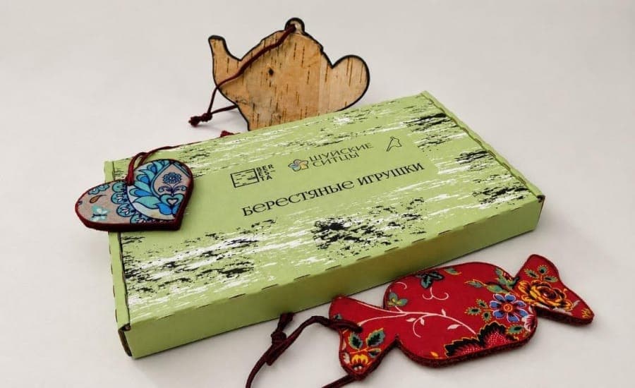 The image shows a light green cardboard box with a distressed, wood-grain texture, featuring a black and white design. The box is adorned with the text "��ересть ��и��иния ��и��иния" in Cyrillic script, which translates to "Handmade gift box". The box is surrounded by three small, handcrafted wooden ornaments. One is a