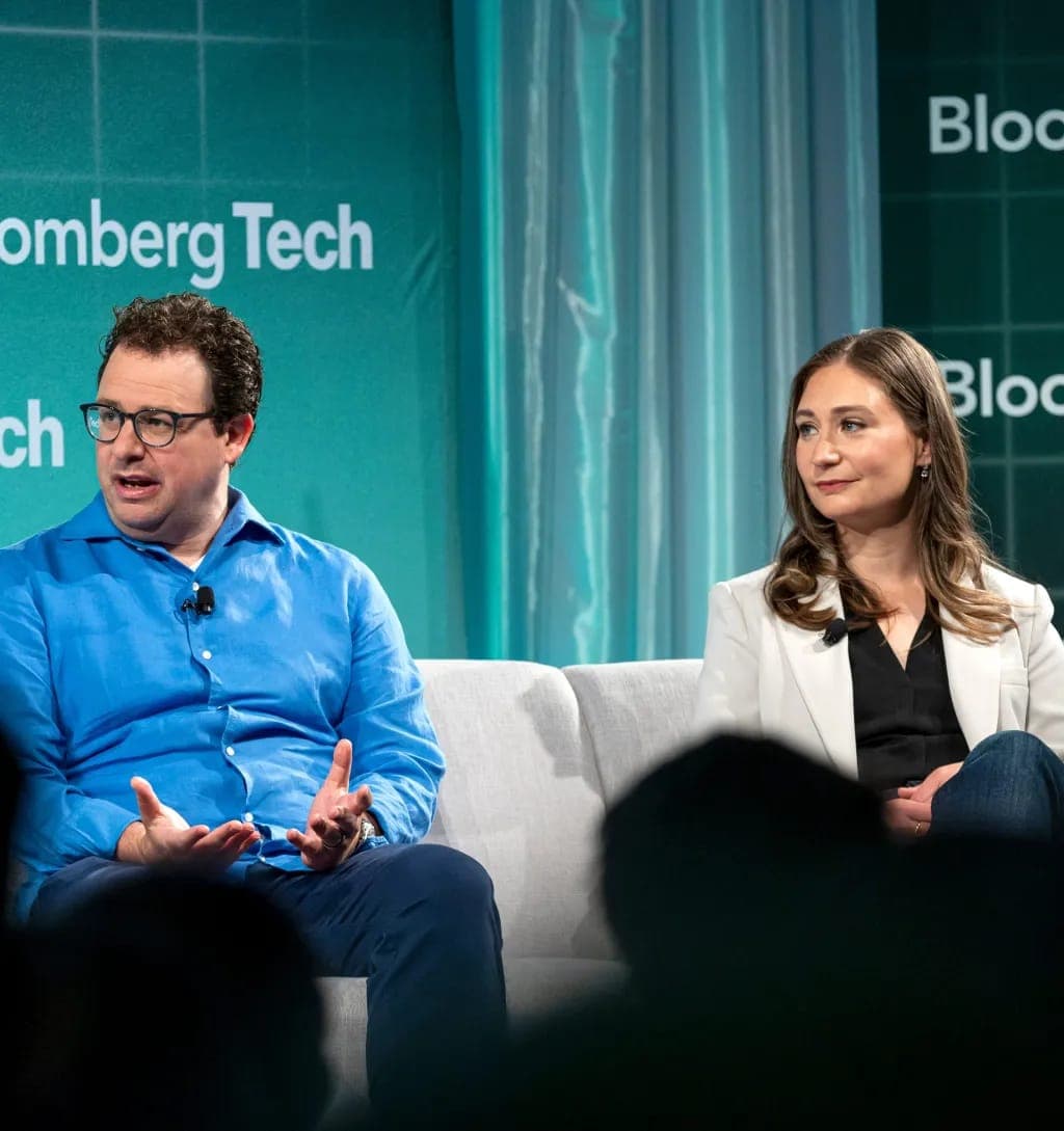 Two people sit on a light gray sofa in front of a teal backdrop with the "Bloomberg Tech" logo. A man on the left in a bright blue shirt and glasses gestures while speaking, and a woman on the right in a white blazer and black top looks toward him.