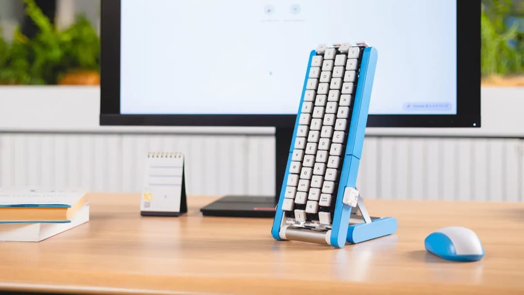 The image shows a workspace with a light brown wooden desk. On the desk, there is a blue and white keyboard stand holding a white keyboard with black keys. A white computer monitor is positioned behind the keyboard stand, displaying a blank white screen. To the left of the keyboard stand, there is a small, white notepa