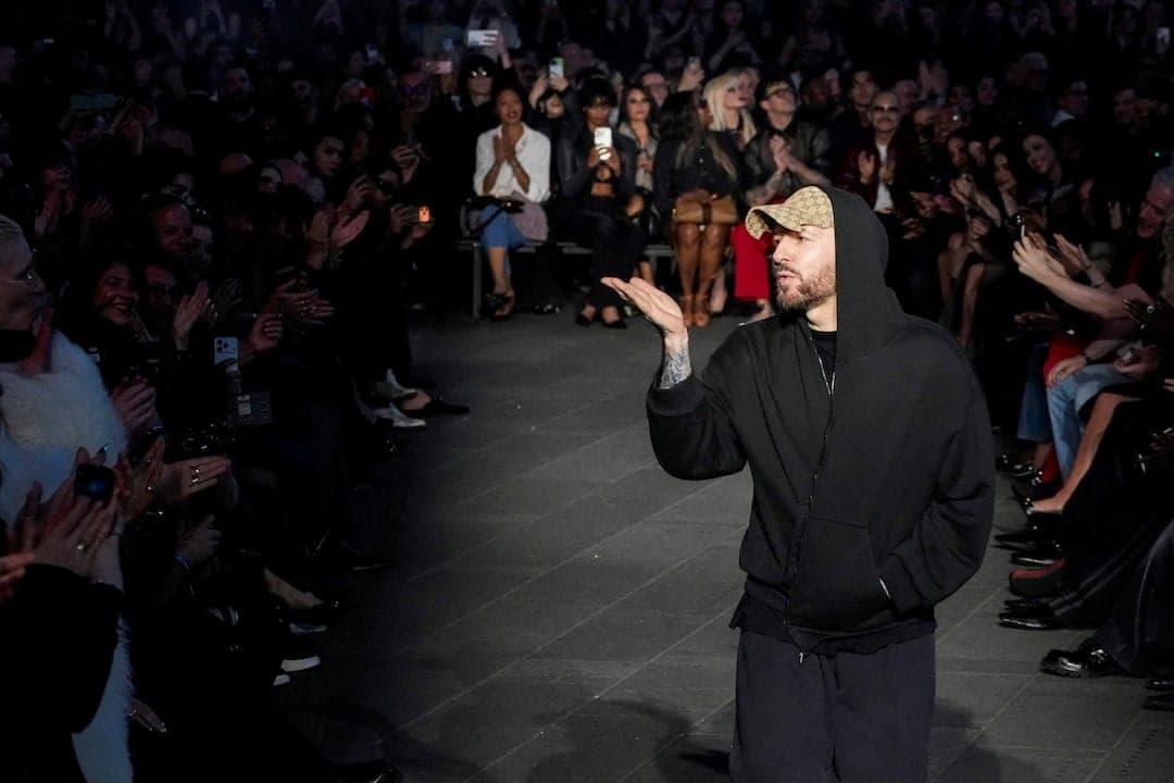 The image captures a fashion show taking place in a dimly lit venue. A male model, wearing a black hoodie and a tan/beige baseball cap, strides confidently down the runway. He is wearing dark-colored pants and has a tattoo on his left arm. The runway is a dark gray or black color, and the audience is seated on either s