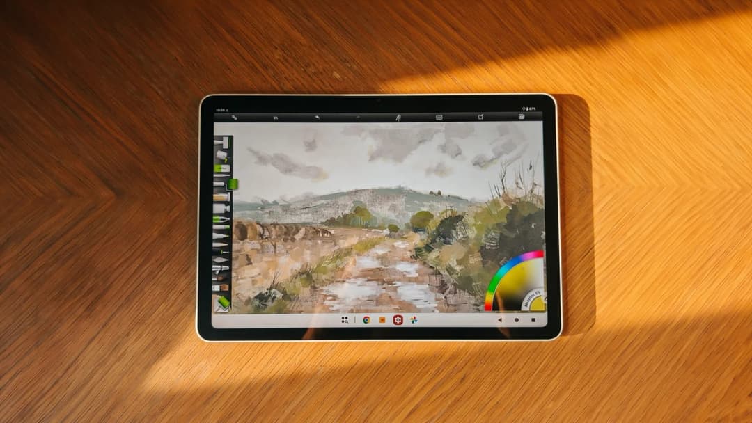 The image shows a tablet computer resting on a wooden surface. The tablet's screen displays a painting of a landscape, featuring a dirt path winding through a field with trees and a hill in the background. The painting is rendered in a style reminiscent of watercolor or impressionism. The tablet's screen also shows a c