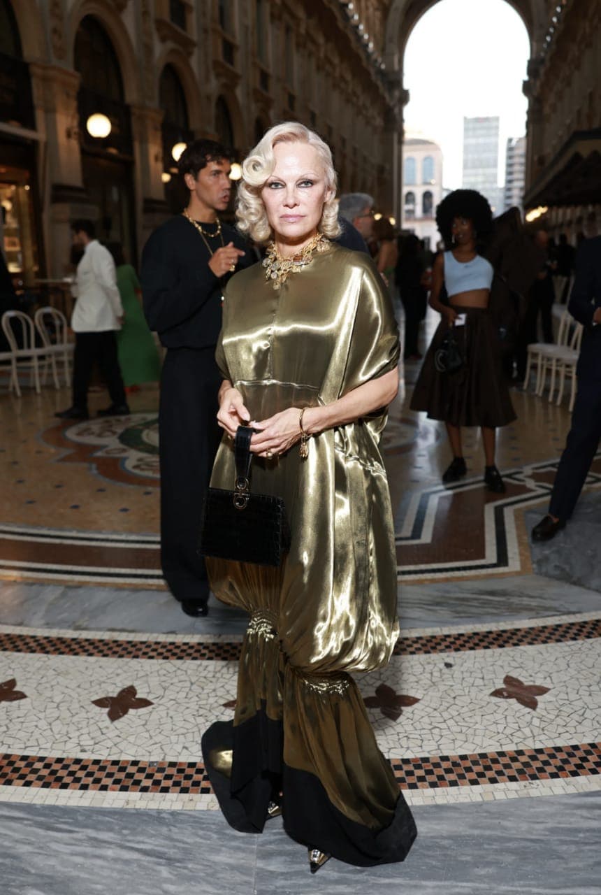 The image shows a woman standing in a grand, ornate hallway. She is dressed in a long, gold dress with a black and gold patterned bottom, and she carries a black purse. Her blonde hair is styled in a vintage, curled fashion. The hallway features a tiled floor with a mosaic pattern of brown, white, and black tiles, and