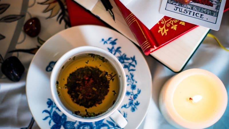 The image shows a white teacup with a blue floral pattern on a matching saucer, filled with a dark brown liquid and a dark red/burgundy tea bag. The teacup and saucer are placed on a white surface, possibly a tablecloth, with a red book and a white card featuring a black and white illustration of a cityscape and the wo
