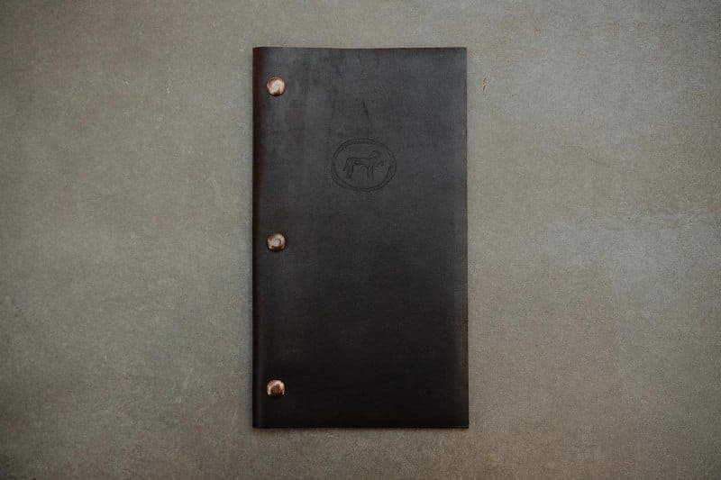 The image shows a black leather menu holder resting on a gray concrete surface. The menu holder has three small, round, metallic-looking fasteners on its sides, and a small, circular logo featuring a stylized animal, possibly a dog or horse, is visible on the front. The menu holder is centrally positioned in the image,