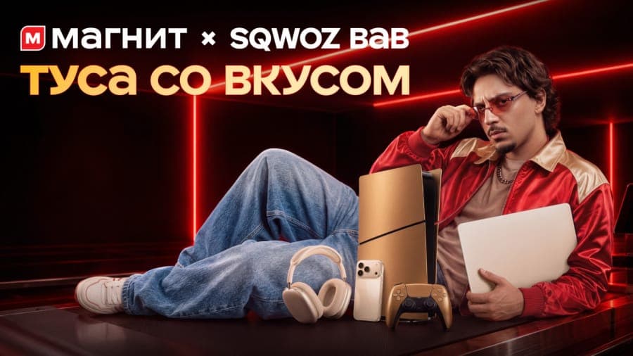The image shows a man reclining on a dark surface, possibly a desk or table, with his legs extended. He is wearing a red jacket, blue jeans, and white sneakers. He is holding a white laptop in his left hand and a white iPhone in his right hand. He is also wearing sunglasses and has a beard. The background is dark with