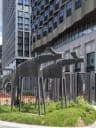 The image shows a modern, multi-story building with a light beige or cream facade and a dark brown or gray roof. The building features a large glass window on the second floor and a smaller one on the first floor. In the foreground, a dark gray metal sculpture of two horses stands on a patch of grass. The sculpture is