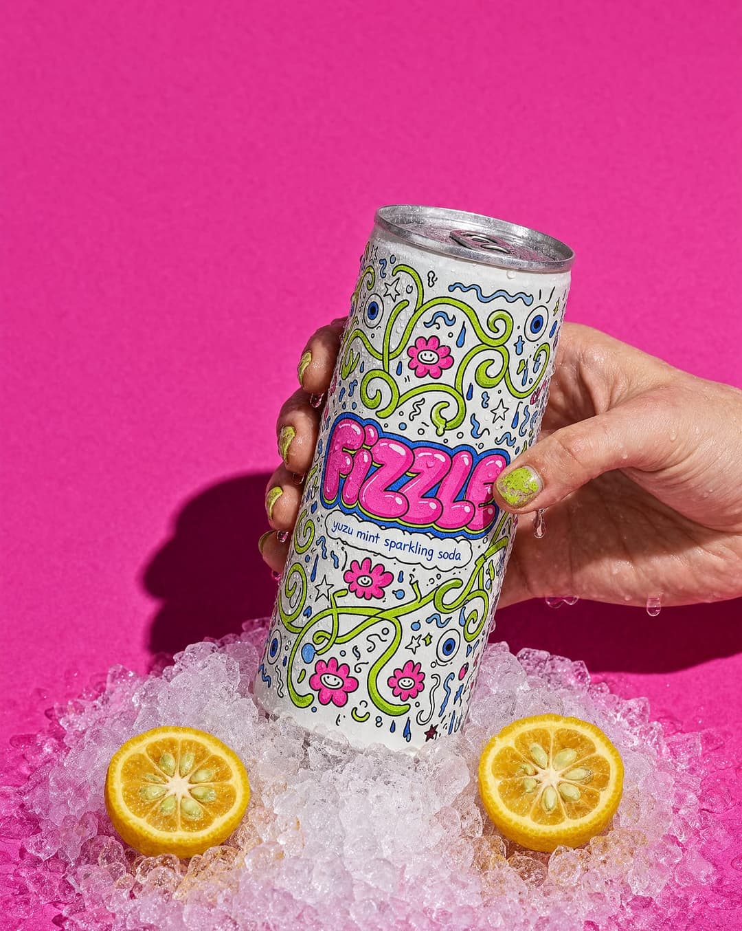 A hand is holding a can of Fizz soda against a vibrant pink background. The can is adorned with a whimsical design featuring pink and green flowers and swirls, along with the word "Fizz" in bold, pink letters. The can is partially covered in ice, and two slices of orange are placed on the ice next to it. The hand has g