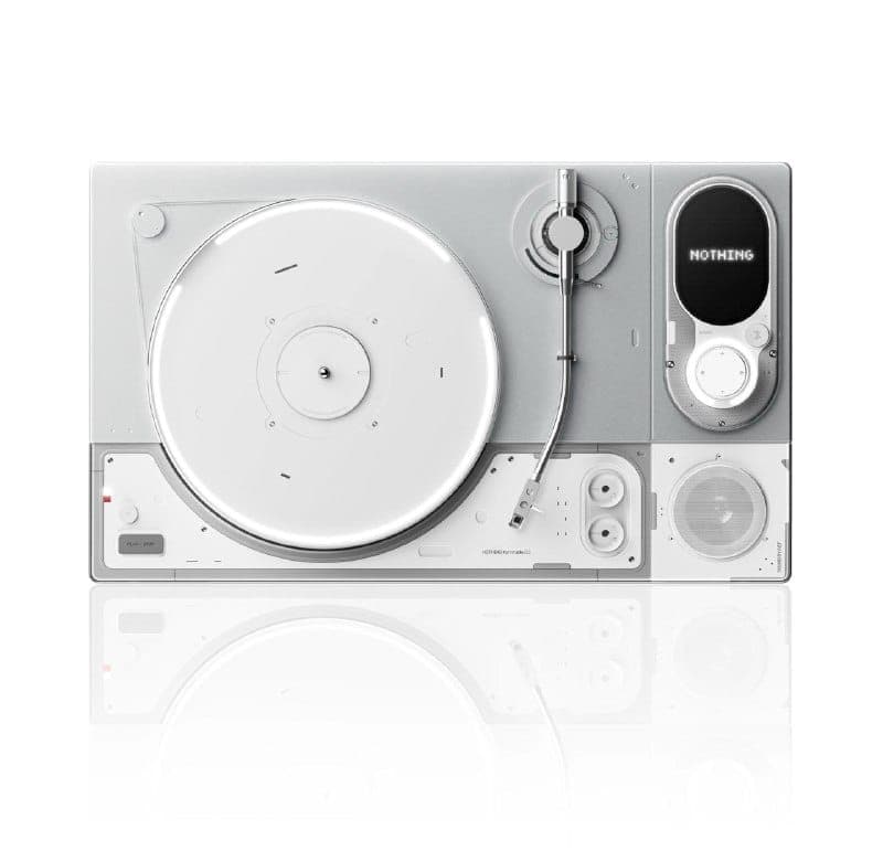 The image presents a sleek, modern, and minimalist device, possibly a DJ controller or music production device. It is predominantly white, with a silver/grey top section that houses a turntable and a small speaker. The turntable is centrally located and has a white circular surface. The device also features a small scr