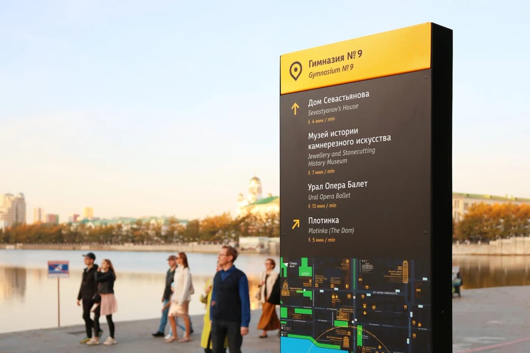 The image shows a tall, rectangular signpost with a black and yellow color scheme, standing on a paved area near a body of water. The signpost is adorned with a map and directional arrows, providing guidance to visitors. The signpost is located near a body of water, with a cityscape visible in the background. Several p