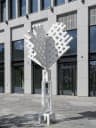 The image shows a white metal sculpture with a heart-like shape, standing on a gray concrete sidewalk in front of a modern gray building with large windows. The sculpture is composed of a series of interconnected white metal pieces, creating a geometric pattern. The building has a flat roof and a series of horizontal l