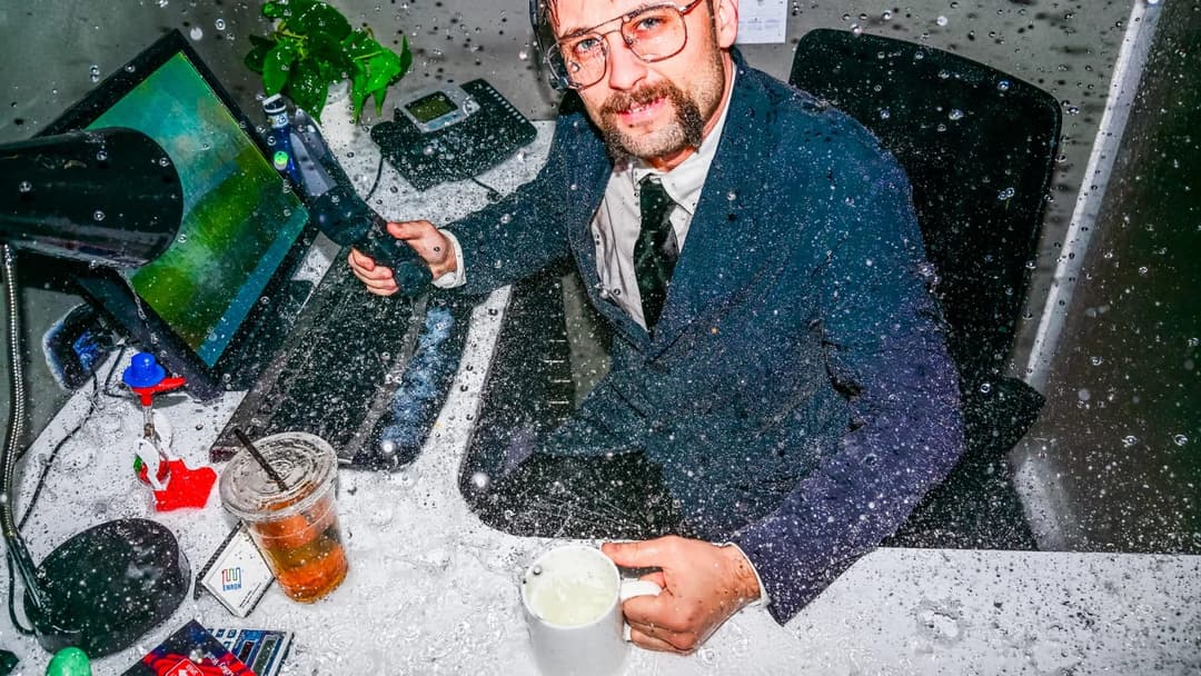 A man in a blue suit and glasses is seated at a desk, holding a black object that appears to be a microphone. He is wearing a dark-colored tie and has a beard. The desk is covered in a layer of white, possibly snow or confetti, giving the scene a festive atmosphere. A computer monitor displays a green landscape, and a