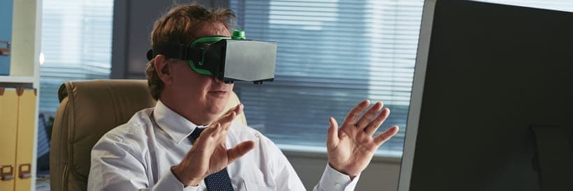 The image shows a man seated in a beige office chair, wearing a white shirt and a dark blue or black tie. He is wearing a black VR headset with a green strap and is gesturing with his hands, suggesting he is engaged in an activity on the computer screen in front of him. The computer monitor is dark gray or black. The b