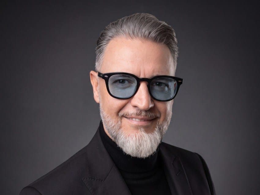 The image shows a man with gray hair and a white beard, wearing a black suit jacket and a black turtleneck. He is wearing black-framed glasses with light blue lenses. The man is looking directly at the camera with a slight smile, and his facial expression is neutral. The background is a solid dark gray color, providing