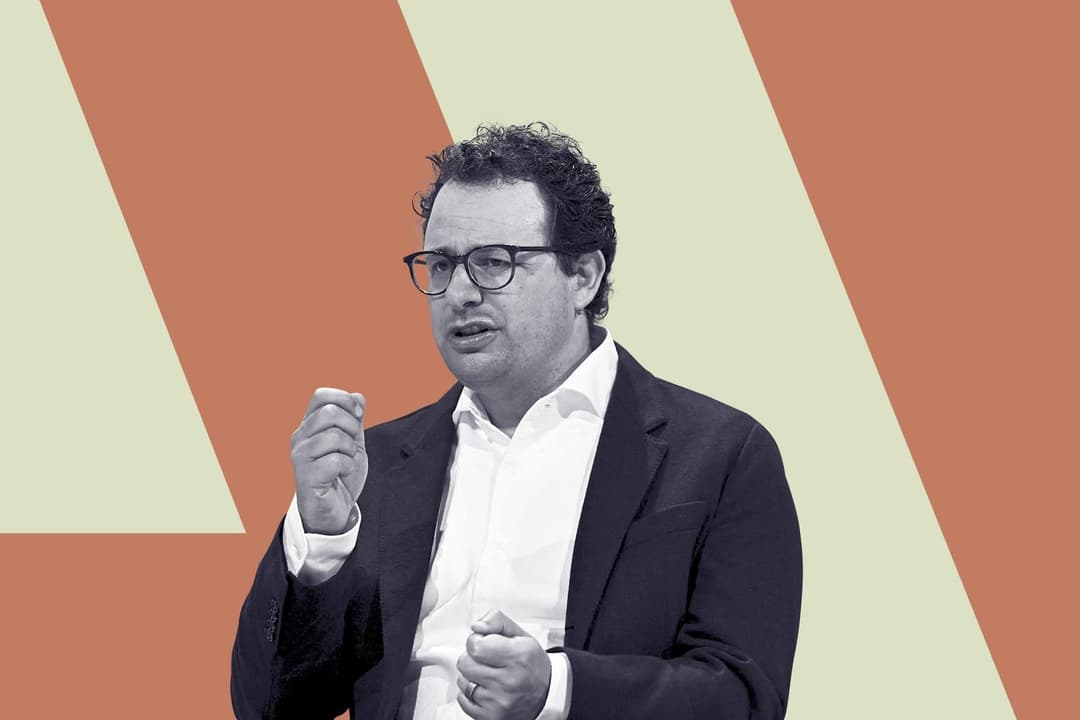 The image shows a man standing against a vibrant orange and beige diagonal stripe background. He is dressed in a black suit and a crisp white shirt, and is wearing glasses. His right hand is raised, and his left hand is clenched into a fist. The man has short, curly hair and a neutral facial expression. The image is in