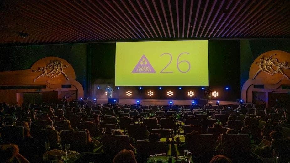The image captures a large, dimly lit auditorium with a high ceiling featuring a wooden slatted design. The walls are adorned with gold-colored sculptures of figures, possibly mythological or mythological in nature. A large screen at the front displays the text "Young Arrows 26", indicating a possible event or presenta