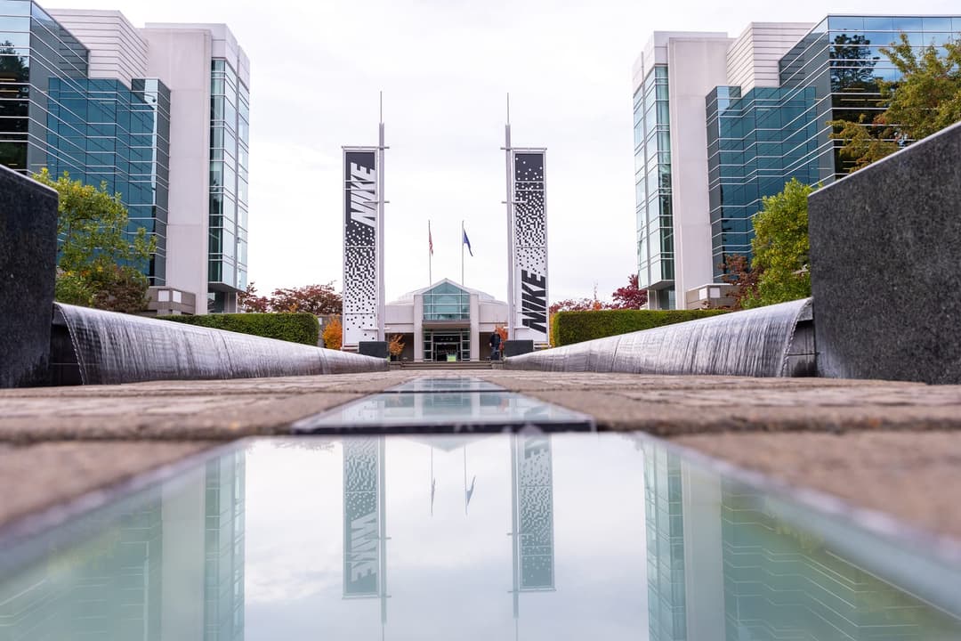 The image captures a view of the Nike headquarters, a modern architectural structure with a glass and steel facade. The building is flanked by two tall, black and white banners with the Nike logo, and the entrance is visible in the center. The perspective is from a low angle, looking up towards the building, creating a