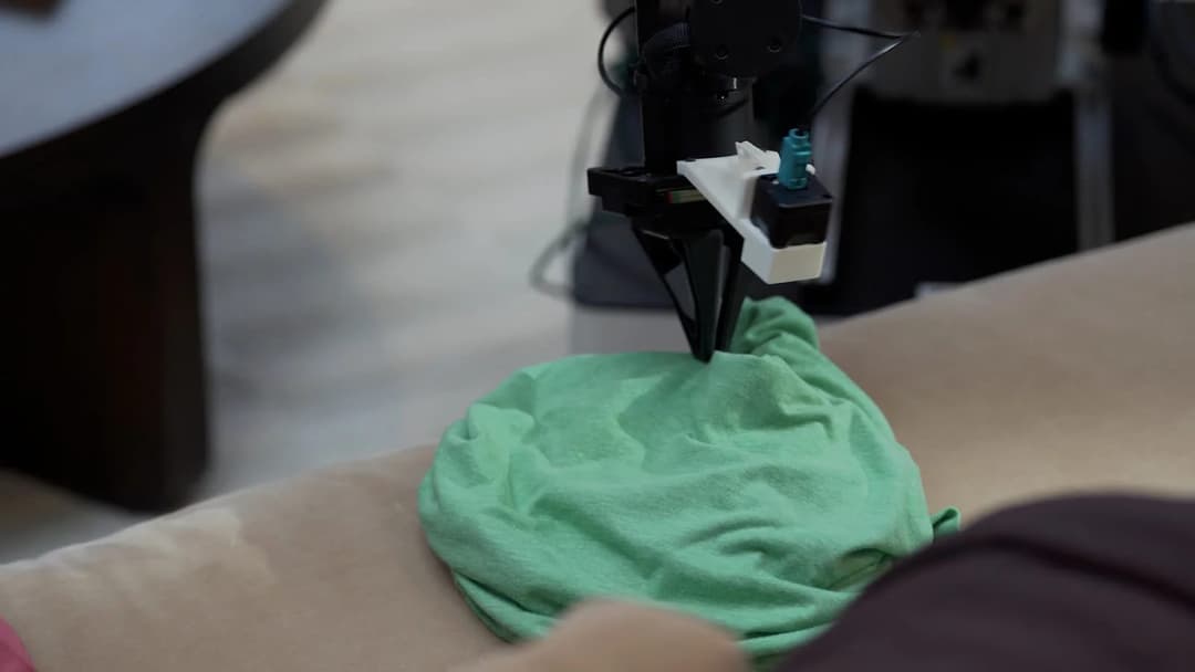 The image shows a black and white robotic arm with a white base and a teal/turquoise colored component, positioned on a beige surface. The arm is holding a green cloth, which appears to be a t-shirt, and is in the process of being sewn onto it. The background is out of focus, but appears to be a room with a wooden tabl