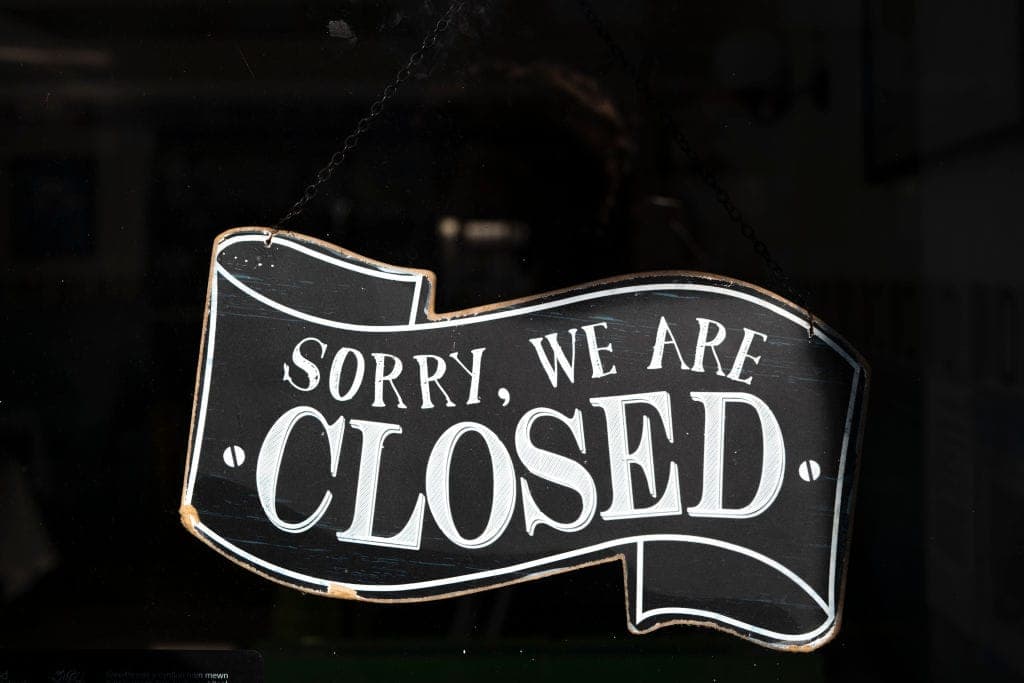 A black ribbon-shaped hanging sign with a white outline and distorted wood-like edges hangs from a chain against a dark window background. The white hand-lettered text reads "SORRY, WE ARE CLOSED"; there is no brand or UI visible.