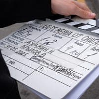The image shows a person's hand holding a black and white film clapperboard. The clapperboard is used to indicate locations on a film set, with lines and numbers written in black ink. The person's hand is positioned on the right side of the clapperboard, suggesting they are in the process of clapping or signaling. The