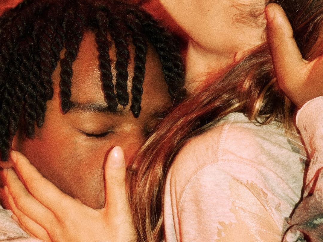 The image shows a close-up of a person's face, with their eyes gently closed, suggesting a moment of rest or sleep. The person has dark brown skin and long, dark hair styled in braids. Their hand is gently resting on their face, and their arm is draped over another person's shoulder. The person receiving the embrace ha