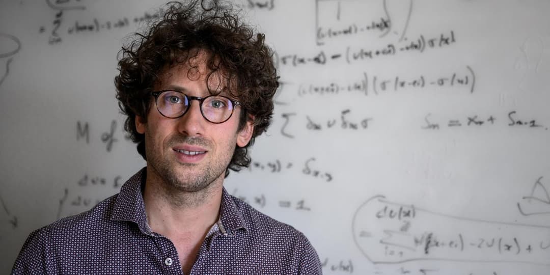 The image shows a man with curly brown hair and glasses standing in front of a whiteboard filled with mathematical equations and diagrams. He is wearing a dark purple or navy blue patterned shirt. The whiteboard is filled with various mathematical equations and diagrams, including some that appear to be related to phys