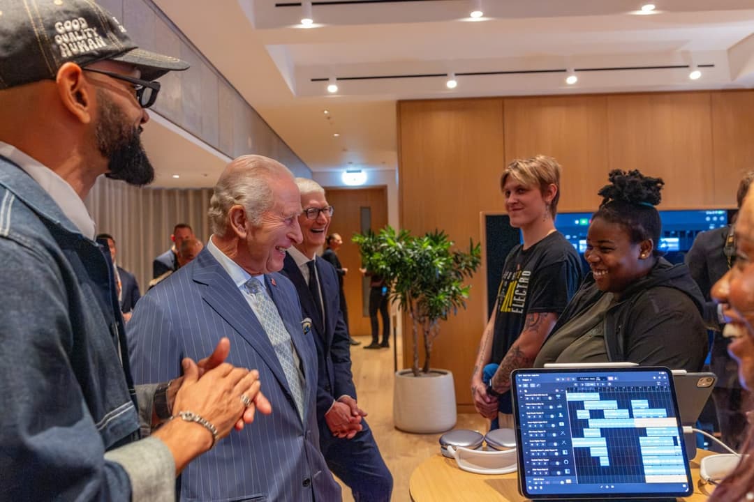 The image shows a group of people gathered in a modern office setting. The room features a wooden wall and ceiling, with a large window providing natural light. A man in a blue suit and a woman in a black t-shirt are engaged in conversation, while others are standing nearby. A man in a blue denim jacket and a woman in
