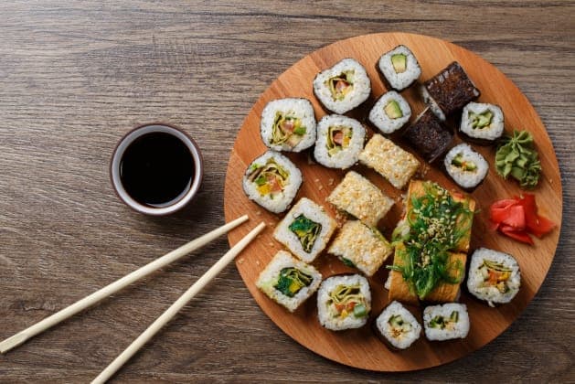 The image shows a wooden table with a round wooden plate holding a variety of sushi rolls. The sushi rolls are arranged in a circular pattern on the plate, with some rolls featuring a variety of toppings, including green seaweed, orange and yellow vegetables, and sesame seeds. A small bowl of dark brown soy sauce is pl
