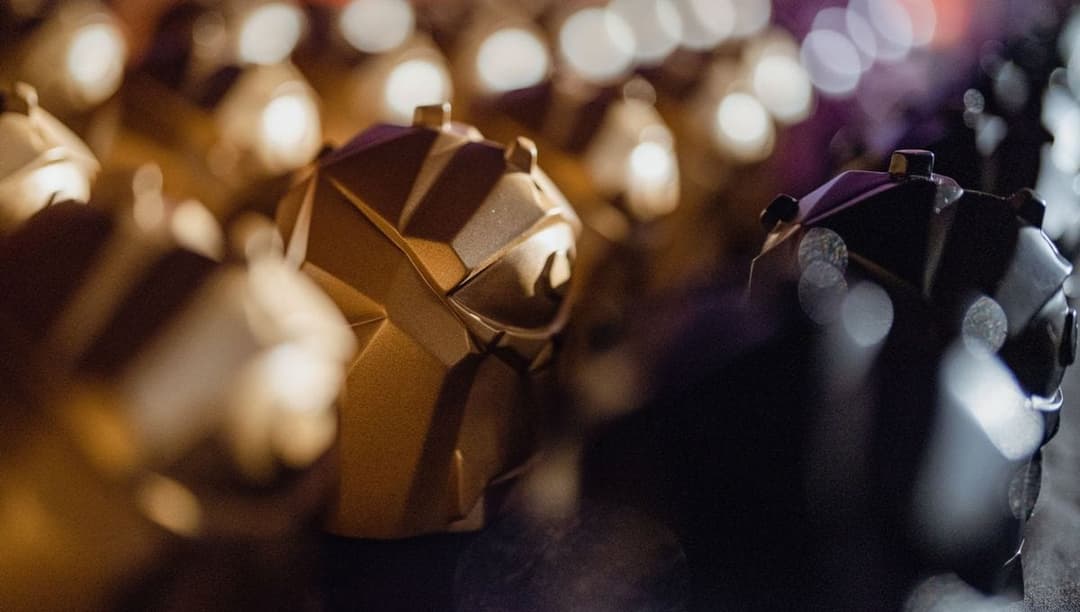 The image shows a close-up view of a collection of golden, geometric objects, possibly sculptures or figurines, arranged in a loose, somewhat haphazard manner. The objects are out of focus, creating a blurred effect that emphasizes their golden hue. The background is a dark, out-of-focus color, possibly black or dark p