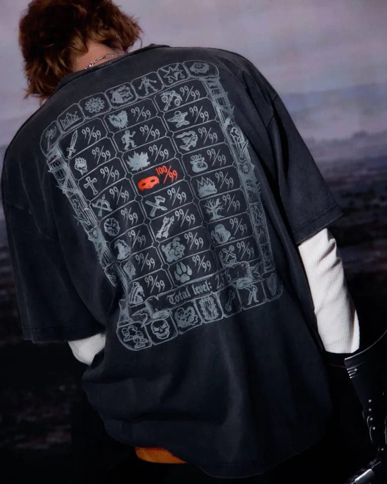 The image shows a person standing against a dark, possibly black, background. The person is wearing a black t-shirt with a unique graphic design on the back. The design features a grid of small, stylized images, possibly representing symbols or artifacts, arranged in a pattern. The design also includes a small, orange,
