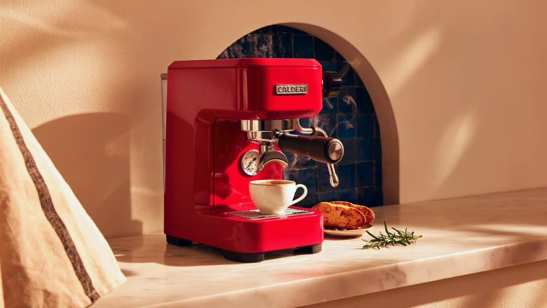 The image shows a red Calder coffee machine with a black handle and a white label on the front, positioned on a white marble countertop. A white coffee cup sits on the countertop, and a small plate with a biscuit or similar baked good is placed next to it. A small sprig of rosemary is also on the countertop. The backdr