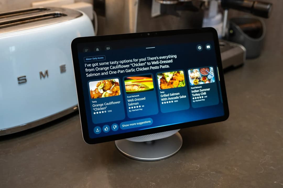 The image shows a kitchen counter with a white Smeg toaster oven and a silver food processor. A tablet computer is positioned on a silver stand in the center of the counter, displaying a menu with a dark blue background and white text. The menu offers a variety of food options, including "Tasty Orange Cauliflower Chick