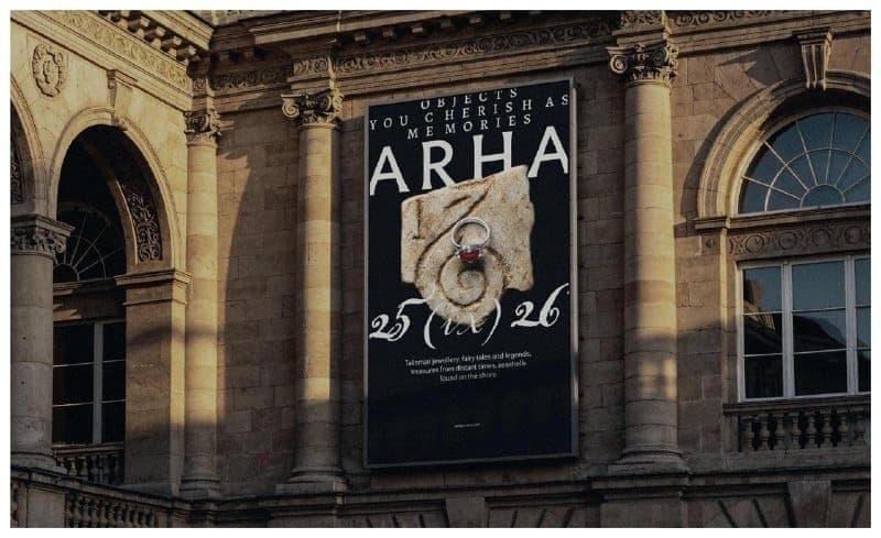 The image shows a large, rectangular black poster affixed to the side of a building. The poster features a white text design that reads "ARHA 25(x)26" and includes a depiction of a stone carving with a ring on it. The building behind the poster is made of light-colored stone and has a classical architectural style, wit