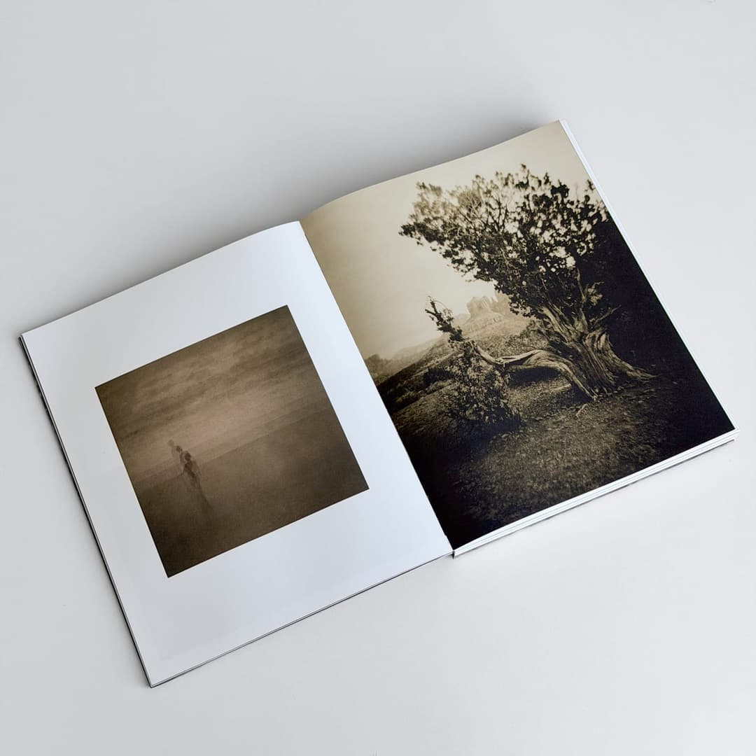 The image shows an open book with two pages, resting on a white surface. The left page features a black and white photograph of a solitary figure standing in a field, with a hazy or misty atmosphere. The right page displays a black and white photograph of a tree with a gnarled trunk and branches, set against a backdrop