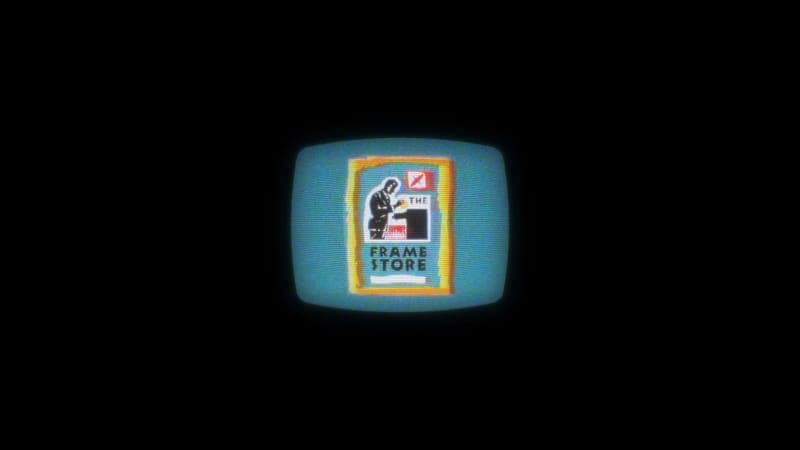 The image shows a vintage television set with a blue screen displaying a logo. The logo features a stylized figure, possibly a man, holding a box or container, and the text "THE FRAME STORE" in white letters. The television is set against a black background, which highlights the vibrant blue screen and the white text o
