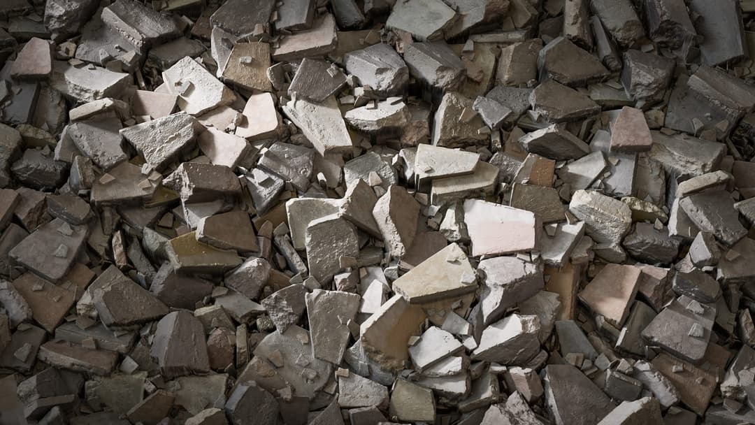 The image shows a pile of broken concrete pieces, varying in shades of gray and brown, scattered haphazardly. The pieces are of different sizes and shapes, with some appearing to be fragments of larger tiles or slabs. The pile is not uniform, with some areas showing a more organized arrangement of pieces. The lighting