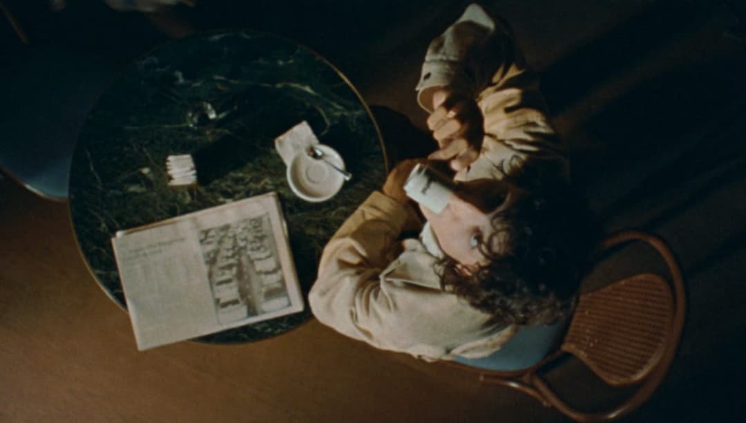 The image shows a person seated at a round, dark green marble table in a dimly lit room. The person is wearing a light beige or cream-colored jacket and is holding a white mug in their right hand. A newspaper is open on the table, and a small white dish with a spoon and a napkin is also present. The person's head is ti