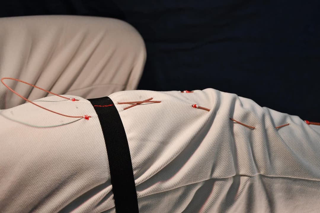 The image shows a close-up view of a person's lower body, likely from the waist down, with a white garment covering the lower half. The garment is adorned with several thin, reddish-brown wires, some of which are connected to a black strap. The wires are attached to the garment in a pattern that suggests they are conne