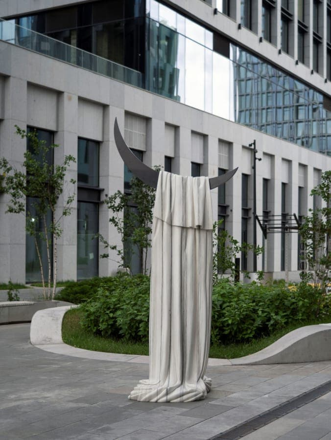 The image shows a large, white, draped sculpture of a crescent moon standing on a gray sidewalk. The sculpture is made of dark metal and is positioned in front of a modern building with a glass facade. The building has a light gray or beige exterior and features a curved or wave-like design element in its facade. The s