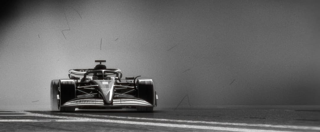 The image depicts a Formula One race car in motion on a wet racetrack. The car, painted in a sleek black and white color scheme, is positioned on the left side of the frame, facing towards the right. The car's body is angled slightly towards the viewer, and the front of the car is pointed towards the right side of the