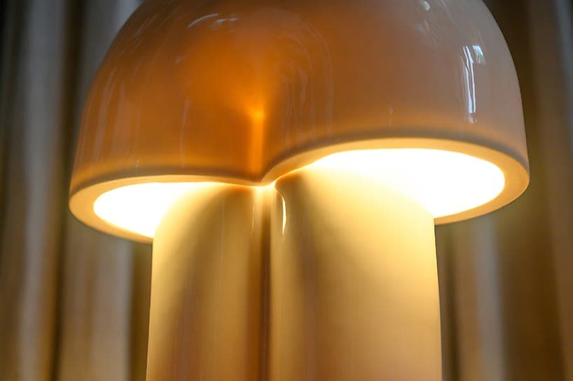 The image shows a close-up view of a lamp with a unique, dome-like shape. The lamp is illuminated, casting a warm, yellowish-orange glow that illuminates the interior. The lamp's base is a light beige or cream color, and the shade is a similar shade of beige or cream. The lamp is positioned against a blurred background