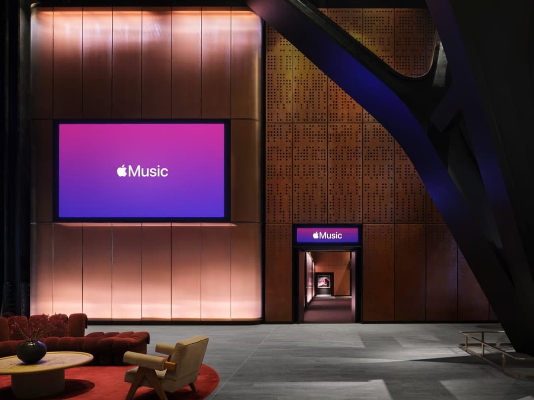 The image depicts a modern, minimalist interior space with a large, rectangular LED screen displaying the Apple logo and the word "Music" in white text on a gradient background of purple and pink. The screen is mounted on a wall with a textured, wood-like surface. To the right of the screen, a dark-colored, curved meta