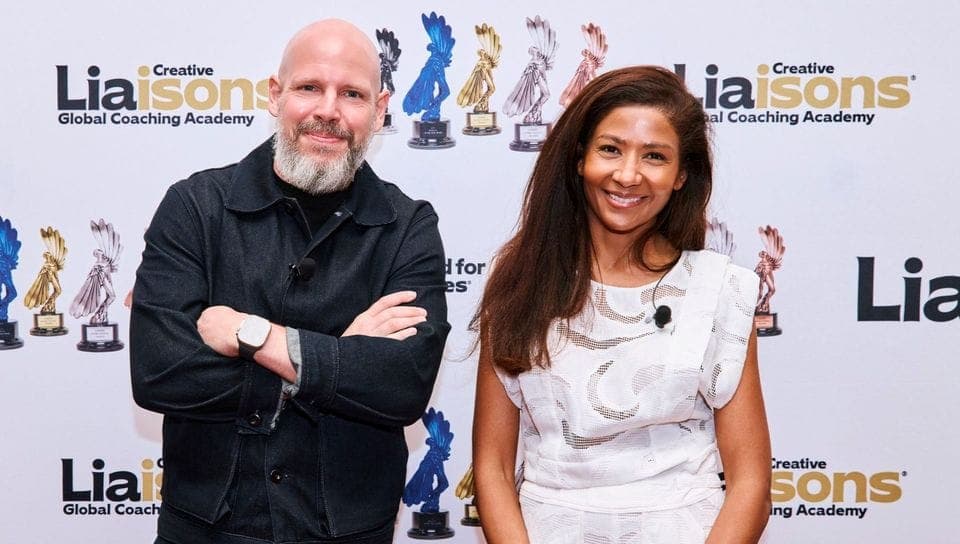 The image shows two individuals standing side by side against a white backdrop adorned with gold and blue text and images of trophies. The man on the left is wearing a black jacket and has a beard, while the woman on the right is wearing a white dress with a patterned design. Both are smiling and looking directly at th