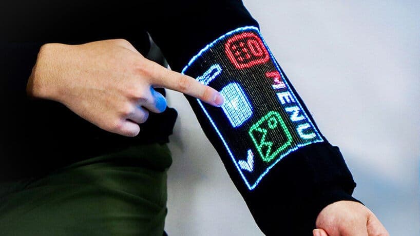 The image shows a person's arm wearing a black sleeve with a digital display. The display features a series of icons, including a red "30" and a green "MENU" icon, along with a white "OK" icon. The person's hand is pointing at the display, suggesting interaction with the technology. The person is wearing a dark-colored